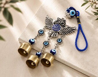 Può includere: Una decorazione da parete con un gufo argentato dalle ali spiegate, decorato con perline di vetro blu a forma di occhio protettivo e tre campanelli in ottone. L'ornamento è sospeso da un cordino blu intrecciato con una perlina a occhio blu.