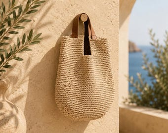 May include: A beige woven straw tote bag with a rounded bottom and two brown leather handles. The bag is suspended from a small wooden peg on a textured light tan wall. The construction features a dense, uniform crochet-like stitch pattern.