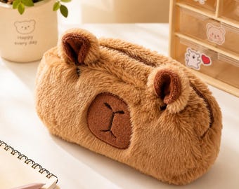 May include: A brown, plush, fuzzy pencil case shaped like a capybara with two small ears on top and a dark brown embroidered nose and mouth detail on the front. The case features a zippered closure along the top edge for storing stationery items.
