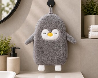 May include: A gray, plush, penguin-shaped hand towel featuring a white face patch, closed eyes, a small yellow beak, and two white feet at the bottom. The towel includes a braided loop at the top for hanging on a black wall-mounted hook.