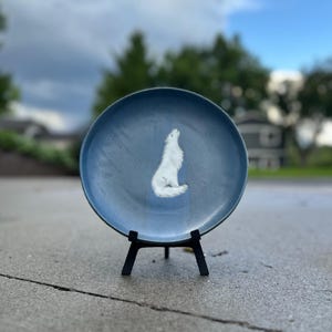 May include: A round, blue ceramic plate featuring a white illustration of a wolf howling at the center. The plate is supported by a black, three-legged display stand, providing a stable base for the circular dish to be viewed upright.
