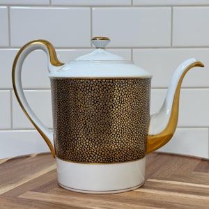 May include: A white porcelain teapot featuring a central band with a black and gold textured pebble pattern. The teapot includes a gold-trimmed handle, a gold-accented spout, and a matching lid with a small gold knob on top, all set against a white base.