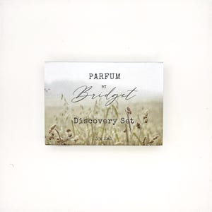 May include: A rectangular perfume discovery set box featuring a soft-focus image of a field of tall grasses. The text on the front reads PARFUM BY Bridget Discovery Set 3 x 2ml in a combination of bold block lettering and elegant script.