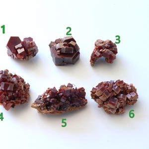 Op de afbeelding: Zes clusters van dieprode, kristallijne vanadinietmineralen gerangschikt in twee rijen. Elk exemplaar heeft hexagonale, tabelvormige kristallen met een glanzende, harsachtige glans. De clusters zijn genummerd van één tot zes in groene tekst.