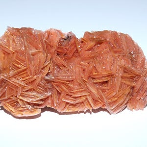 May include: A raw mineral specimen featuring a cluster of interlocking, blade-like crystals in a salmon-pink to reddish-orange hue. The structure is dense and irregular, composed of numerous thin, sharp-edged crystalline formations.