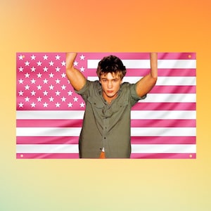May include: A rectangular wall banner featuring a pink and white American flag pattern. Centered in front of the flag is a photograph of a person with short brown hair wearing a short-sleeved olive green button-down shirt with two chest pockets.