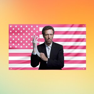 Könnte beinhalten: Eine rechteckige Wandflagge mit dem Porträt eines Mannes im schwarzen Anzug und weißem Hemd, der einen weißen Latexhandschuh anzieht. Der Hintergrund der Flagge ist eine rosa-weiße Version der amerikanischen Flagge mit Sternen und Streifen.