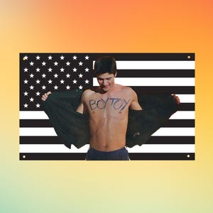 Op de afbeelding: Een rechthoekige muurvlag met een zwart-wit Amerikaans vlagpatroon met sterren en strepen. In het midden staat een foto van een persoon met donker haar, in een donkere korte broek, die een donker jasje opent om de tekst BOY TOY op de borst te tonen.