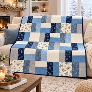 Peut inclure: Une courtepointe rectangulaire présentant un motif en patchwork composé de blocs de tissu alternés dans des tons bleu marine, bleu clair, gris et crème. Les motifs incluent des flocons de neige blancs, des aiguilles de pin et des unis.