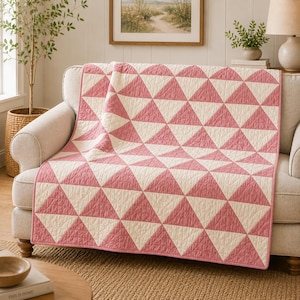 Puede incluir: Una colcha rectangular con un patrón geométrico repetitivo de triángulos alternos en color rosa palo y blanco roto. La tela presenta una textura acolchada visible en toda la superficie y está rematada con un borde de tela rosa sólido.