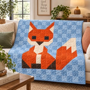 Op de afbeelding: Een gewatteerde plaid met een gepixeld vossenontwerp in oranje, wit en donkerbruin tegen een lichtblauwe geblokte achtergrond. De vos heeft puntige oren, een witte borst en een grote pluizige staart, opgebouwd uit kleine vierkante lapjes stof.
