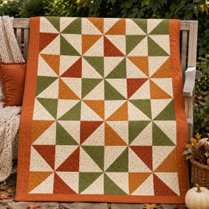 May include: A rectangular quilt featuring a repeating geometric pattern of half-square triangles in shades of cream, olive green, burnt orange, and rust. The quilt is finished with a solid burnt orange fabric border along all four edges.