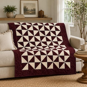 Puede incluir: Una colcha rectangular con un patrón repetitivo de molinillos en tela color burdeos oscuro y crema. La colcha tiene un borde burdeos sólido con una textura acolchada sutil y está colocada sobre un sofá de color claro con un cojín crema.