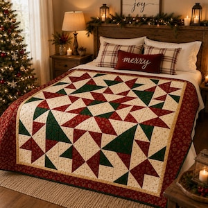 Op de afbeelding: Een quilt met een geometrisch sterpatroon in rood, donkergroen en crème. De quilt heeft een brede rode rand met een subtiele bloemenprint en ligt over een bed met witte kussens en een rechthoekig rood kussen met het woord merry erop.