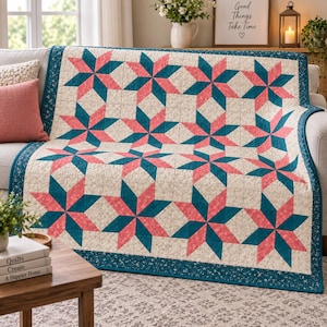Puede incluir: Una colcha con un patrón repetitivo de estrellas en tonos rosa coral y azul cerceta profundo sobre un fondo color crema con un sutil estampado floral. La colcha está rematada con un borde azul cerceta con un pequeño diseño floral blanco.
