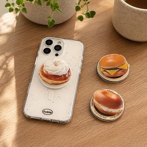 May include: A clear smartphone case with black speckles and the word lucky printed at the bottom, featuring a doughnut-shaped phone grip. Two additional circular phone grips are nearby, one shaped like a cheeseburger and one like a bagel.