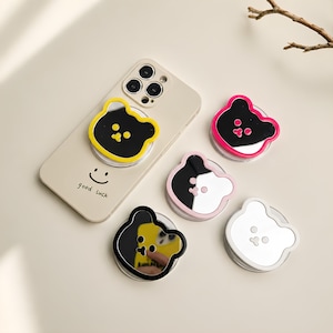 Könnte beinhalten: Eine beige Smartphone-Hülle mit einem Smiley-Gesicht und dem Text good luck, inklusive eines gelben, bärenförmigen Spiegel-Handyhalters. Vier weitere bärenförmige Spiegel-Handyhalter sind in den Randfarben Pink, Schwarz, Hellrosa und Weiß enthalten.
