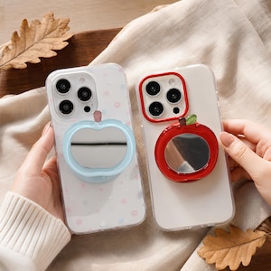 May include: Two clear smartphone cases featuring integrated apple-shaped mirrors on the back. One case has a light blue apple mirror and a star-patterned back, while the other has a solid red apple mirror with a green stem and a plain back.