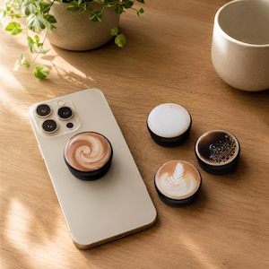 Peut inclure: Un smartphone de couleur claire avec un support de téléphone circulaire arborant un motif de tourbillon de café brun et blanc fixé au dos. Trois autres supports circulaires sont à proximité, montrant de la mousse, du café noir et du latte art.