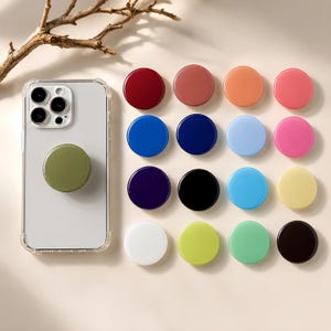 Puede incluir: Una funda transparente para smartphone con un soporte circular de color verde oliva, acompañada de una colección de dieciséis soportes circulares adicionales en varios colores sólidos, incluyendo rojo, azul, rosa, amarillo, negro y blanco.