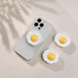 May include: Three phone grips shaped like fried eggs, each featuring a glossy white irregular border and a raised, smooth yellow center. One grip is attached to the back of a light gray smartphone, while two additional grips are placed nearby.