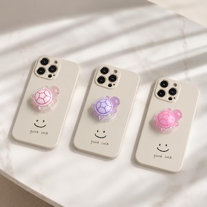 May include: Three beige smartphone cases, each featuring a black smiley face, the text "good luck," and a small, translucent turtle-shaped phone grip. The turtles are coloured in shades of pink and purple, attached to the centre of each case.