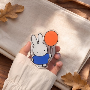 May include: A clear acrylic phone grip featuring a cartoon rabbit character with long white ears, a blue shirt, and a black 'x' mouth, holding a solid orange circular balloon. The design is printed on a transparent base with a circular mount.