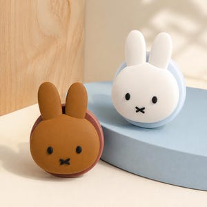 May include: Two circular decorative items featuring rabbit faces with long ears, black oval eyes, and a black cross-shaped mouth. One item is brown with a dark brown base, and the other is white with a light blue base, both having smooth surfaces.
