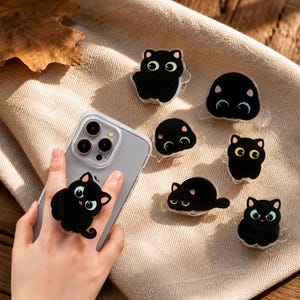 May include: A clear smartphone case with a black cat phone grip attached, accompanied by six additional black cat phone grips. Each grip features a cartoon-style black cat with large, expressive eyes and small pink ears, designed for mobile devices.
