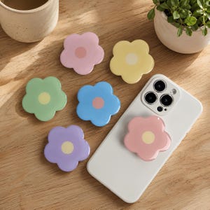 May include: Six flower-shaped phone grips in pastel colors including pink, yellow, light green, blue, and purple. Each grip features a contrasting circular center. One pink flower grip is attached to the back of a white smartphone.