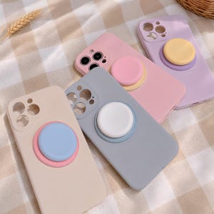 Peut inclure: Quatre coques de smartphone aux couleurs pastel, notamment beige, gris, rose et lavande. Chaque coque présente un support de préhension circulaire et pliable à l'arrière dans une couleur contrastante. Les coques ont des découpes pour un appareil photo à trois objectifs.