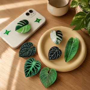 May include: A collection of seven leaf-shaped phone grips featuring various botanical patterns, including variegated monstera, dark veined foliage, and bright green textures. One grip is attached to a white smartphone with green star decals.