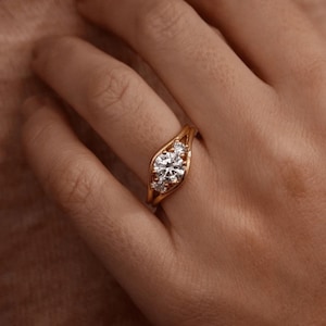 Peut inclure: Une bague en or ornée d'une pierre précieuse transparente ronde centrale, entourée de deux pierres plus petites de taille ronde. L'anneau présente un design divisé qui s'incurve autour des pierres, créant un style croisé poli.