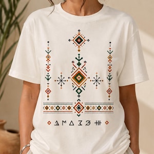 May include: A cream-colored short-sleeve t-shirt featuring a central graphic design composed of geometric shapes, diamonds, and star-like patterns in shades of dark green, burnt orange, and black, with the text "AMAZIGH" printed at the bottom.