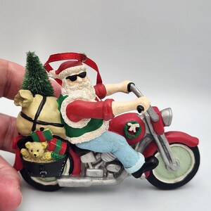 Puede incluir: Un adorno de Santa Claus montado en una motocicleta roja, con gafas de sol, chaleco rojo y verde y pantalones azules. Lleva un saco beige, un pequeño árbol verde y una cesta con un oso de peluche y regalos envueltos atrás.