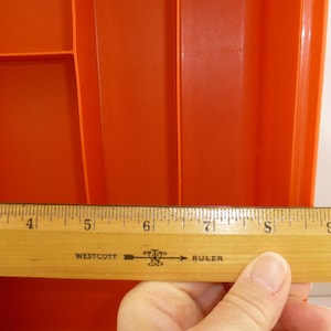 May include: A bright orange plastic drawer organizer tray with multiple compartments. A wooden Westcott ruler is positioned horizontally across the front, showing a measurement scale from three to ten inches.
