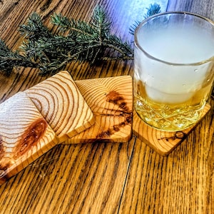 Peut inclure: Un ensemble de quatre sous-verres carrés en bois présentant des motifs de grain naturel visibles et des nœuds sombres. Un sous-verre soutient un verre transparent rempli d'un liquide doré et de glace, avec une branche de pin verte à proximité.