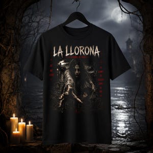 May include: A black short-sleeve t-shirt featuring a graphic print of a ghostly figure reaching forward. The text reads LA LLORONA, LA LEYENDA DE MARIA, AY MIS HIJOS, and SU LLANTO SE ESCUCHA JUNTO AL RIO in white and red lettering on the front.
