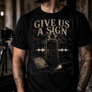 May include: A black short-sleeved t-shirt featuring a graphic print with the text GIVE US A SIGN and LISTEN CLOSELY. The design includes a digital voice recorder, a field notes notebook, a flashlight, and a sound wave against a dark background.