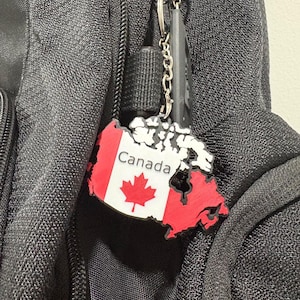 Könnte beinhalten: Ein Schlüsselanhänger in Form der Landkarte von Kanada mit einem weißen Zentrum, einem roten Ahornblatt und dem in schwarzer Schrift gedruckten Wort Canada, flankiert von roten Abschnitten auf der linken und rechten Seite, an einem silbernen Ring.
