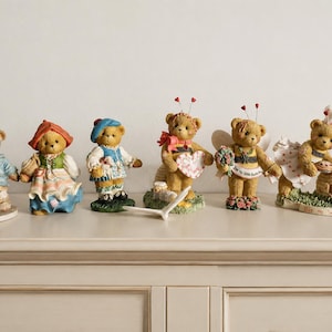 Puede incluir: Una colección de seis figuras decorativas de osos. Los osos llevan varios atuendos, incluyendo una chaqueta azul, una capucha roja, una boina azul, un disfraz de abeja con un corazón, un disfraz de abeja con alas y un traje de chef con colmena.