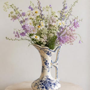 May include: A cream-coloured ceramic pitcher vase featuring a blue floral pattern and a decorative handle, filled with a vibrant arrangement of white daisies, purple bellflowers, and delicate green wildflowers, creating a tall, textured display.