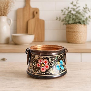 Peut inclure: Un petit pot rond en cuivre avec un extérieur sombre et patiné orné de motifs floraux peints à la main en rouge, bleu et blanc. Le récipient possède deux anses latérales en anneau métallique et un bord intérieur en cuivre poli.
