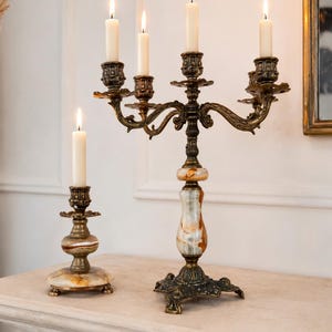 May include: A five-arm brass candelabra and a single-arm brass candlestick, both featuring ornate metal detailing and polished stone bases with swirling cream and brown patterns. Each candle holder supports a single lit white taper candle.