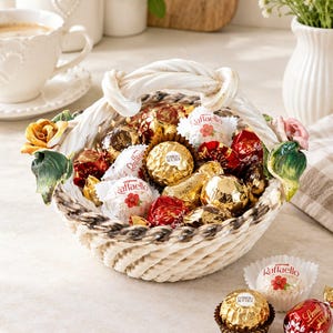 Könnte beinhalten: Ein Keramikkorb mit geflochtenem Henkel und dekorativen Keramikrosen an den Seiten, gefüllt mit einer Auswahl an einzeln verpackten Pralinen, darunter goldfolierte Ferrero Rocher und weiß verpackte Raffaello-Konfekte.