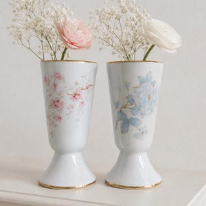 Może przedstawiać: Dwie białe porcelanowe wazony ze złoconymi brzegami i podstawami. Jeden wazon ma różowy wzór kwiatowy, drugi niebieski. Każdy wazon zawiera białą gipsówkę oraz pojedynczy kwiat jaskra w kolorze różowym lub białym.