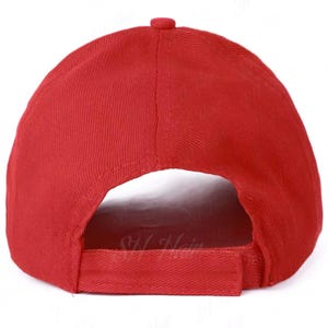 May include: A solid red baseball cap featuring a structured crown with stitched panels and a button at the top. The back of the cap includes an open arch design and an adjustable fabric strap with a hook and loop fastener for a secure fit.