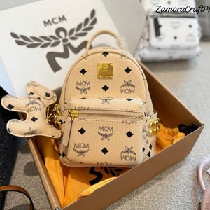 以下が含まれることがあります： 黒いMCMロゴパターン、前面のゴールドトーンのメタルプレート、ジッパー付きフロントポケット、黒いダイヤモンドのアクセントが付いたベージュのウサギ型バッグチャームが特徴のベージュのミニバックパック。段ボール箱に入った状態で提示されています。
