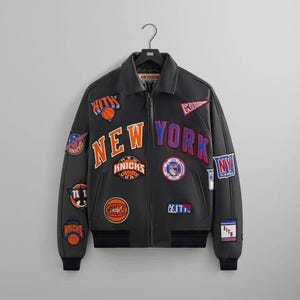 May include: A black leather bomber jacket featuring a full-length front zip, a collar, and ribbed cuffs. The jacket is decorated with various embroidered patches, including the text NEW YORK in orange and blue, KITH logos, and basketball motifs.