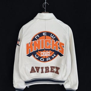 May include: An off-white varsity jacket featuring a large orange and navy blue New York Knicks basketball logo on the back with the text KITH inside. The word AVIREX is embroidered below in navy blue. A small American flag patch is on the sleeve.